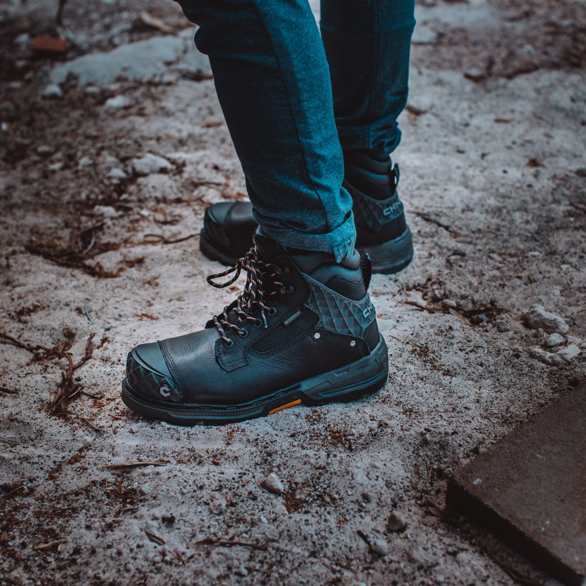 Pallet Jack 6" Comp Toe Men’s Waterproof Work Boot in Black, worn by a person standing on a rough, dirt-covered surface. The boots feature a tough leather exterior, diamond-patterned reinforcement on the heel and toe, and sturdy laces, emphasizing their rugged durability and readiness for demanding conditions.