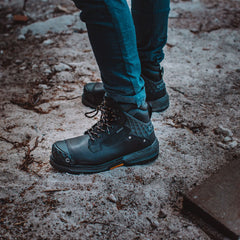 Pallet Jack 6" Comp Toe Men’s Waterproof Work Boot in Black, worn by a person standing on a rough, dirt-covered surface. The boots feature a tough leather exterior, diamond-patterned reinforcement on the heel and toe, and sturdy laces, emphasizing their rugged durability and readiness for demanding conditions.