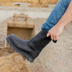 A worker is pulling on the black Boss 11” Comp Toe Men’s Work Boot while standing on construction machinery, showcasing the boot's durable design and tough performance for demanding work environments. The textured sole and reinforced toe highlight its strength and stability.