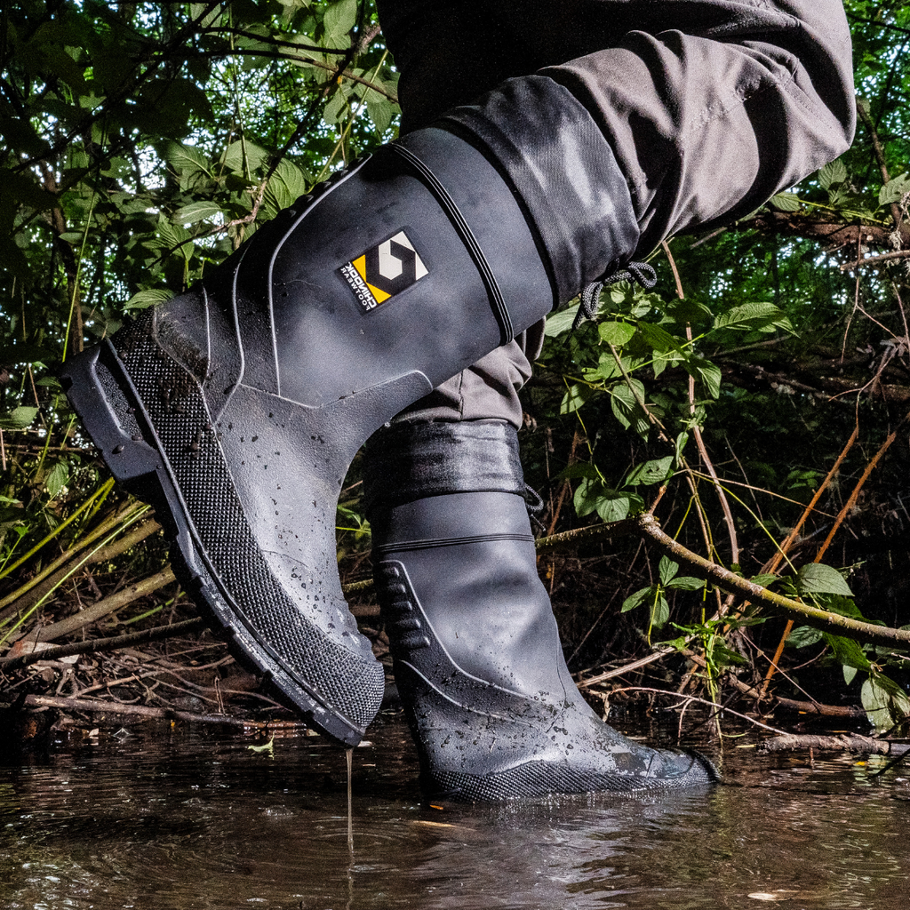 A men wearing Badaxe Regrind Waterproof Rubber Boots stands in a shallow stream, with water droplets on the boots. The rugged black boots feature a textured sole and reinforced design, ideal for wet, muddy environments and tough outdoor conditions.