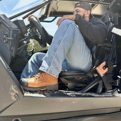 A man wearing Maverick 6” Comp Toe Men’s Waterproof Work Boots in wheat color relaxes in an off-road vehicle, with one foot casually propped on the side rail. These rugged boots, designed with reinforced toes, are built to handle it all—whether you're tackling tough outdoor terrain or powering through a long day on the job.