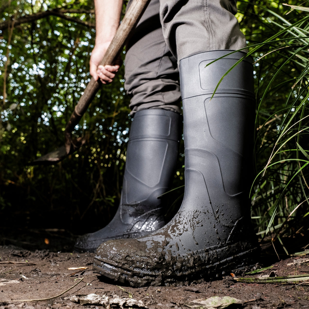 A man wearing Kickaxe Regrind 15" Men's Waterproof Rubber Boots stands in a muddy forest, holding a shovel. The boots, coated in mud, highlight their durable, waterproof design, built for tough outdoor work and wet conditions.