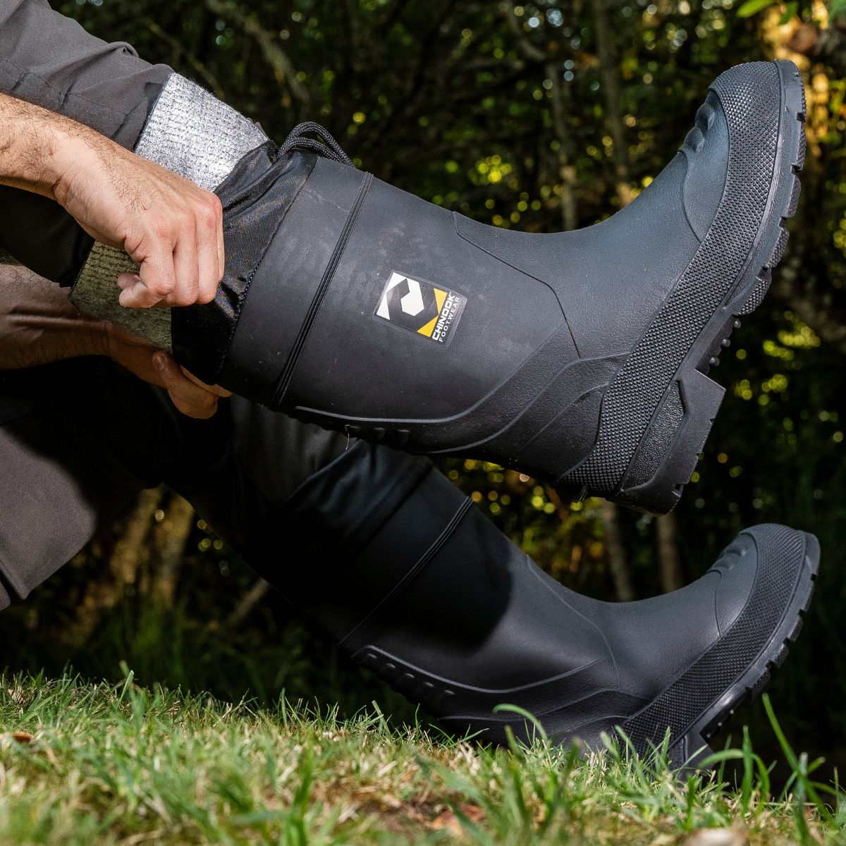 A men adjusting their Badaxe Regrind Waterproof Rubber Boot in black while sitting on grass. The boots feature a rugged design with a textured sole and reinforced areas, built for durability and protection in wet, muddy environments.