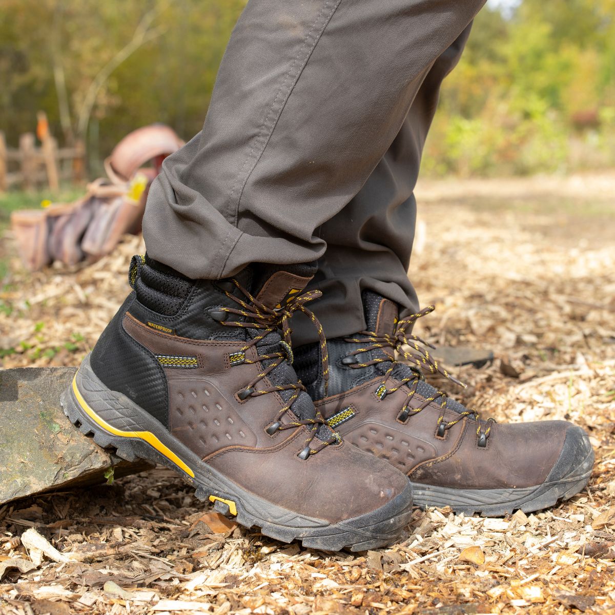 A close-up of a hiker wearing the Olympic 6" Comp Toe Men’s Waterproof Hiker Boot in Brown. The boots feature a rugged design with reinforced laces and a sturdy outsole, ideal for outdoor exploration. The natural outdoor setting emphasizes the boots' durability and comfort, while the waterproof construction ensures reliable protection in varying weather conditions. The boots offer a secure fit, making them perfect for long hikes and adventurous trails.