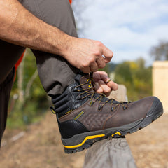 A hiker tightens the laces of the Olympic 6" Comp Toe Men’s Waterproof Hiker Boot in Brown. The boot features a rugged design with reinforced stitching and a durable outsole, perfect for outdoor exploration. The hiker is outdoors, with a natural backdrop, highlighting the boot’s comfort and reliability in various weather conditions. Its waterproof construction and secure fit make it an ideal choice for extended hikes and outdoor activities.