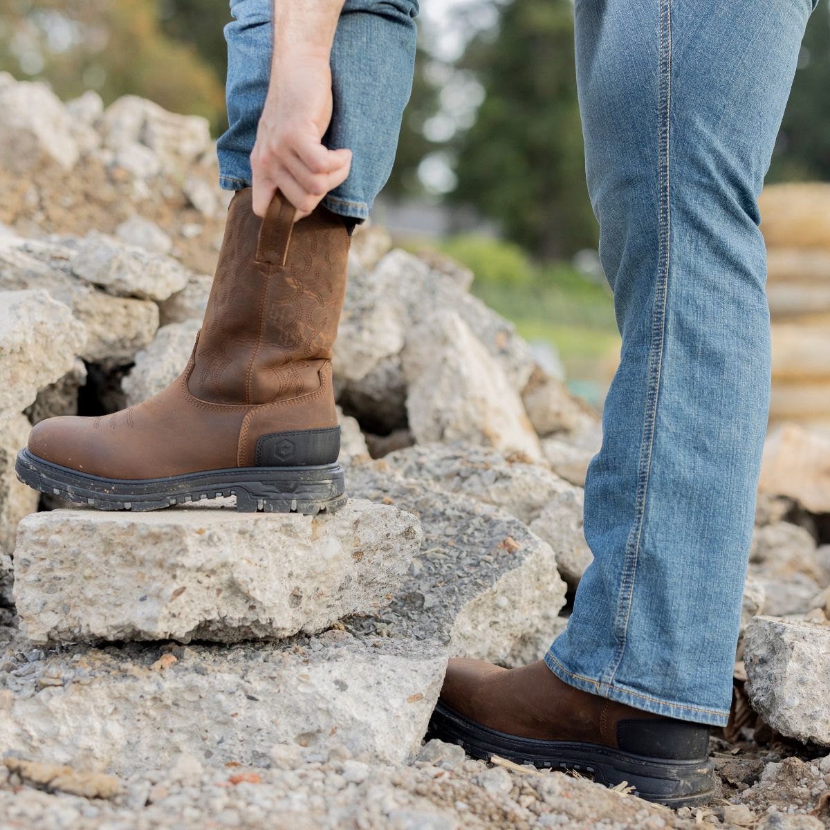 A man pulling on the Trailboss 11” Soft Toe Men’s Work Boot while standing on rocky terrain. The boot features a durable leather design with pull tabs and a rugged outsole, ideal for tough outdoor and work environments.